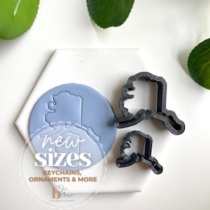 May include: Two black cookie cutters in the shape of the state of Alaska. The cutters are on a white surface with a blue background showing the outline of Alaska. The text "new sizes" is written in black on the blue background. The text "keychains, ornaments & more" is written in black below the blue background.