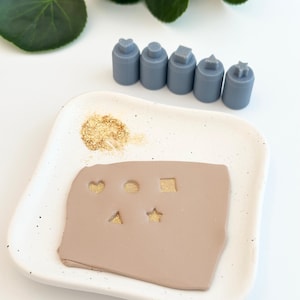 May include: A set of five grey polymer clay stamps in the shapes of a heart, circle, square, triangle, and star. The stamps are arranged in a row on a white surface. A small dish with a brown clay surface and gold glitter is in the foreground. The stamps have been used to create the shapes in the clay.