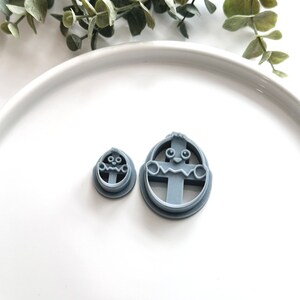 May include: Two gray plastic cookie cutters shaped like Easter eggs with a cartoon face. The larger cutter has a cross-shaped design inside the egg.