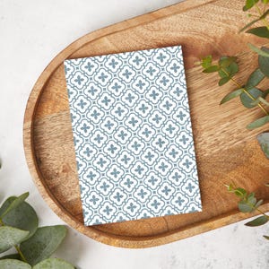 Puede incluir: Una tarjeta rectangular con un patrón repetitivo de cuadrifolios azul grisáceo sobre un fondo blanco. La tarjeta descansa sobre una bandeja de madera con acabado natural, rodeada de follaje verde.