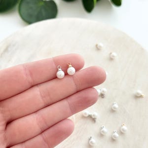 May include: Close-up of small, white pearl charms with silver and gold-colored findings. Several charms are scattered on a white surface. The charms are round and have a delicate, elegant appearance, suitable for jewelry making.