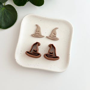May include: Four witch hat-shaped items in two different shades of brown sit on a speckled white ceramic plate. These are likely Halloween-themed craft or decorative items.