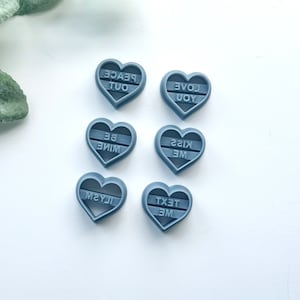 May include: Six gray heart-shaped cookie cutters with different phrases: "Peace Out", "Love You", "Be Mine", "Kiss Me", "Text Me", and "Miss You".