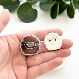 May include: A gray plastic cookie cutter shaped like a sheep with a white fondant sheep next to it.