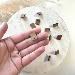 May include: A set of small, square wooden earrings with a natural wood finish. The earrings have a silver post and are displayed on a wooden surface.