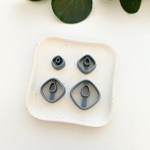 May include: Four gray plastic cookie cutters in the shape of a diamond with a smaller circle inside. The cutters are on a white speckled surface.