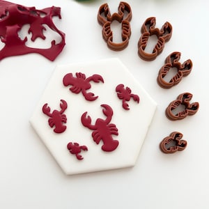 May include: A collection of red lobster-shaped fondant decorations and brown lobster-shaped cookie cutters. The fondant lobsters are arranged on a white hexagonal surface. Several brown plastic cookie cutters are also visible.