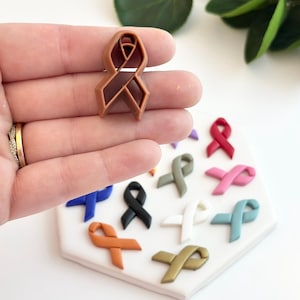 Puede incluir: Surtido de dijes de cinta de colores en varios colores, incluyendo azul, negro, naranja, blanco, dorado, rosa y verde azulado. Los dijes están dispuestos en un círculo sobre una superficie blanca.