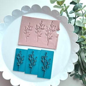 May include: Three blue silicone stamps with a leafy branch design. The stamps are on a white plate with a scalloped edge. A pink silicone stamp with the same design is in the background.