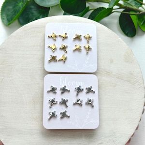 May include: Two cards with a white background and the word "bloom" printed on them. Each card has a set of frog-shaped earrings. One set is gold and the other is silver.