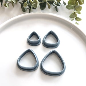 May include: Four gray plastic cookie cutters shaped like teardrops, arranged on a white surface. The cutters are different sizes, with the largest cutter at the bottom of the image.