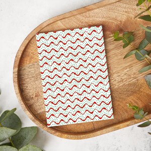 May include: A rectangular card with a white background featuring a repeating pattern of red wavy lines and small green dots. The card is placed on a wooden tray with a natural finish, with green foliage in the corners.