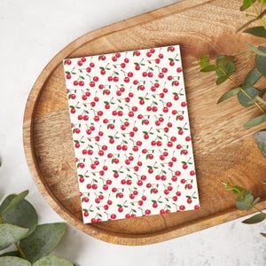 May include: A white card with a repeating pattern of red cherries on stems. The card is on a wooden tray.