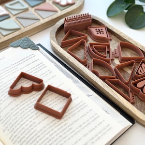 May include: A collection of terracotta-colored polymer clay cutters in various shapes, including triangles, squares, and heart shapes, displayed on a wooden tray. A book with a decorative bookmark is in the foreground. The word "bloom" is visible on some cutters.