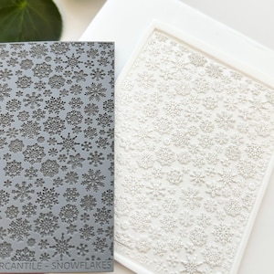 Puede incluir: Un sello de silicona gris con un patrón de copos de nieve. El sello está etiquetado como "Cantile - Snowflakes".