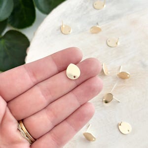 May include: Gold teardrop shaped earring blanks with a hole in the center. The earring blanks are on a wooden surface.