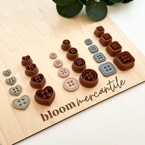 May include: An assortment of brown, grey, and pink button-shaped cookie cutters displayed on a light wood board. The cutters include heart, square, and round shapes. The board has the text "bloom mercantile" engraved on it.