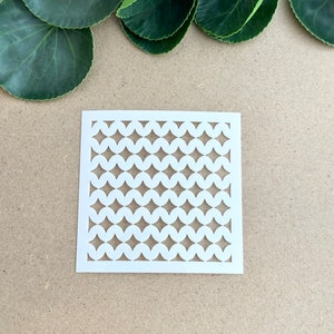 May include: White stencil with a repeating pattern of circles and diamonds. The stencil is on a brown surface.