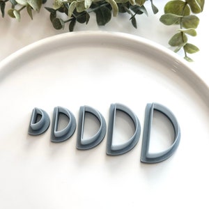 Puede incluir: Conjunto de cinco cortadores de galletas de plástico gris en forma de semicírculos, dispuestos en orden de tamaño de menor a mayor.