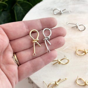 Pode incluir: Dois pares de brincos em forma de nó, um par é dourado e o outro é prateado. Os brincos são exibidos em uma superfície branca.