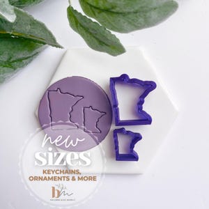 May include: Three purple plastic cookie cutters in the shape of the state of Minnesota. The cutters are on a white surface. The text "new sizes" is written in black and white on a purple background. The text "keychains, ornaments & more" is written in black and white on a purple background. The text "bh" is written in black and white on a purple background.