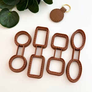 Könnte beinhalten: Satz mit vier braunen Plastik-Ausstechern in Form eines Kreises, Rechtecks, Quadrats und Ovals. Jeder Ausstecher hat eine genähte Kante. Ein brauner Leder-Schlüsselanhänger mit einem goldenen Ring ist ebenfalls enthalten.
