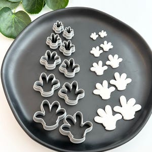 May include: A set of 14 cookie cutters in the shape of a hand with four fingers. The cutters are made of metal and are arranged in two rows on a black plate. The cutters are in various sizes, with the largest cutters at the bottom of the image and the smallest cutters at the top.