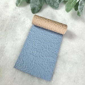 May include: A close-up of a blue textured leather sheet rolled up with a tan textured leather sheet. The blue sheet has a bumpy, uneven surface.