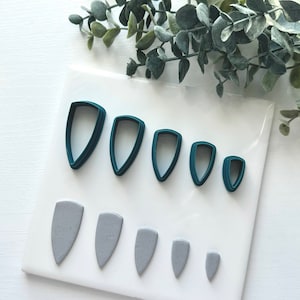 Może przedstawiać: Zestaw sześciu tealowych plastikowych foremek do cięcia w różnych rozmiarach, w kształcie spiczastego owalu, oraz sześć szarych form z gliny wykonanych za pomocą foremek.