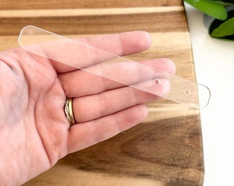 Puede incluir: Una regla de plástico transparente con dos agujeros en la parte superior. La regla mide aproximadamente 15 cm de largo.