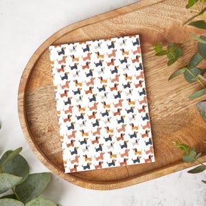Puede incluir: Un cuaderno blanco con un patrón repetido de perros salchicha coloridos. Los perros están en varias poses, incluyendo sentados, de pie y corriendo. El cuaderno es perfecto para tomar notas, escribir ideas o llevar un diario.