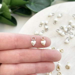 May include: Close-up of heart-shaped pearl charms. Two charms are held between fingers, with many more scattered in the background. The charms have a silver or gold-coloured ring at the top and a small pearl at the bottom.