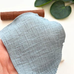 May include: A piece of light blue polymer clay with a textured, woven appearance is held in a hand. A brown clay roller is in the background. The clay has a rough, fabric-like texture. The image is well-lit, showing the details of the clay.