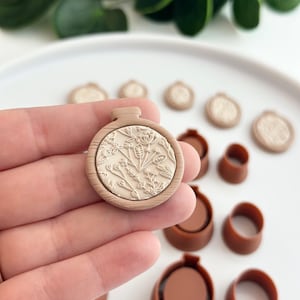 May include: A wooden embroidery hoop with a floral design. The hoop is beige and the design is white. The hoop is surrounded by other crafting supplies.
