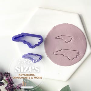 May include: Two purple plastic cookie cutters in the shape of the state of North Carolina. The cutters are on a white surface with a pink circle of clay showing the outline of the state. The text "new sizes, keychains, ornaments & more" is below the cutters.