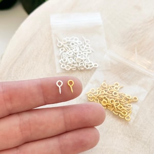 Puede incluir: Dos bolsas de pequeñas alfileres de ojo dorados y plateados. Los alfileres de ojo se muestran en una imagen de primer plano con un dedo sosteniendo dos de los alfileres.