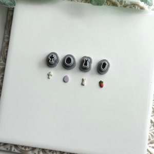 May include: A set of six small grey plastic cookie cutters in the shapes of a cross, circle, bunny, dress, and two different acorns. The cutters are arranged on a white tile surface.