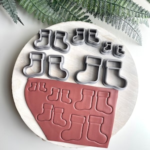 Puede incluir: Un juego de nueve cortadores de galletas de metal con forma de calcetines de Navidad, con una hoja de fondant rojo que muestra las formas recortadas.
