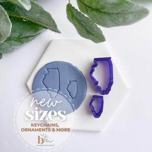 May include: Two purple plastic cookie cutters in the shape of the state of Illinois. The cutters are on a white hexagonal tile with a blue clay imprint of the state of Illinois. The text "new sizes" and "keychains, ornaments & more" is printed on the tile.