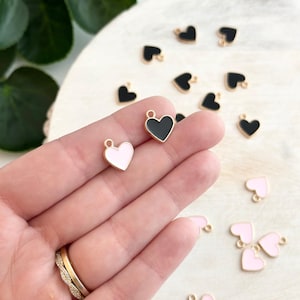 May include: A close-up of a hand holding two gold heart charms. One charm is black with a gold outline and the other is pink with a gold outline. There are other charms scattered on a white surface.