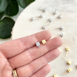 Könnte beinhalten: Nahaufnahme von zwei quadratischen Ohrring-Verschlüssen, einer silberfarben und einer goldfarben, mit einem Sprungring, der oben befestigt ist. Die Verschlüsse befinden sich auf einer Hand mit einem Ring am Finger.