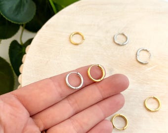 Op de afbeelding: Een close-up van een hand die twee kleine zilveren oorringen vasthoudt. De oorringen liggen op een houten oppervlak met andere oorringen in goud en zilver.