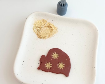 May include: A white ceramic plate with a red clay ornament shaped like a mitten. The mitten has three gold glitter stars on it. There is a pile of gold glitter on the plate.