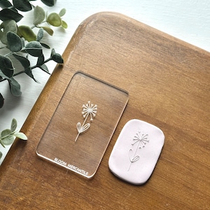 Puede incluir: Un sello de acrílico transparente con un dibujo lineal simple de una flor. El sello está sobre una superficie de madera y tiene el texto "BLOOM MERCANTILE" en la parte inferior.