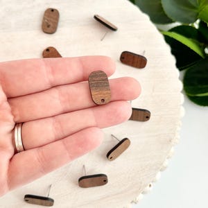 May include: A close-up of a hand holding a wooden oval earring with a dark brown stain. The earring has a small hole at the top for a post. There are several other wooden earrings on a white surface in the background.