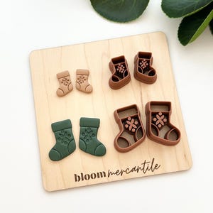 May include: A wooden board displays six Christmas stocking cookie cutters in various shades of brown and green. The cutters are arranged in pairs, with each stocking featuring a snowflake design. The text "bloom mercantile" is printed on the board.