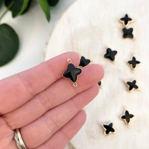 Puede incluir: Encantos de tréboles negros de cuatro hojas con bordes y lazos dorados. Los encantos están esparcidos sobre una superficie de color claro. Un encanto se sostiene entre los dedos. Los encantos son probablemente para la fabricación de joyas u otras manualidades.