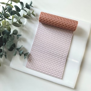 May include: A pink silicone rolling pin with a textured surface, resembling fish scales, is shown on a white surface. The rolling pin is rolled up and the textured surface is visible.