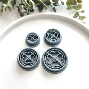 May include: Four gray plastic cookie cutters with a four-leaf clover design. The cutters are arranged in a row on a white surface.
