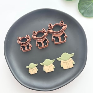 Puede incluir: Un juego de seis cortadores de galletas con la forma de un personaje de una popular franquicia de ciencia ficción. Los cortadores están dispuestos sobre un plato gris oscuro. Tres son marrones y tres son verdes y beige.